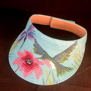 May include: A blue visor with a painted design of a hummingbird in flight, a pink flower, and a dragonfly. The visor has a light pink adjustable strap.