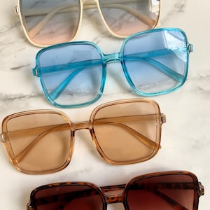 May include: Four pairs of square-framed sunglasses in various colors. The top pair has a cream-colored frame with blue and pink gradient lenses. The second pair has a light blue frame and blue lenses. The third pair has a brown frame and lenses. The bottom pair has a tortoiseshell frame and brown lenses.