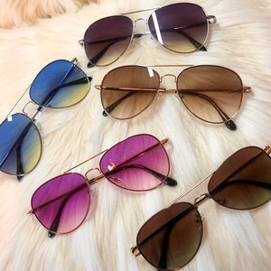 May include: Five pairs of aviator sunglasses with gradient lenses. The sunglasses have gold frames and different colored lenses: purple, brown, blue, and two pairs of brown.