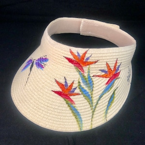 May include: A white straw sun visor with a painted design of tropical flowers and a dragonfly. The flowers are red, orange, yellow, blue, and green.
