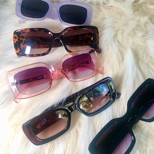 May include: Four pairs of sunglasses with different colored frames and lenses. The sunglasses are arranged in a row on a white fuzzy surface. The sunglasses are all rectangular in shape and have a modern style.