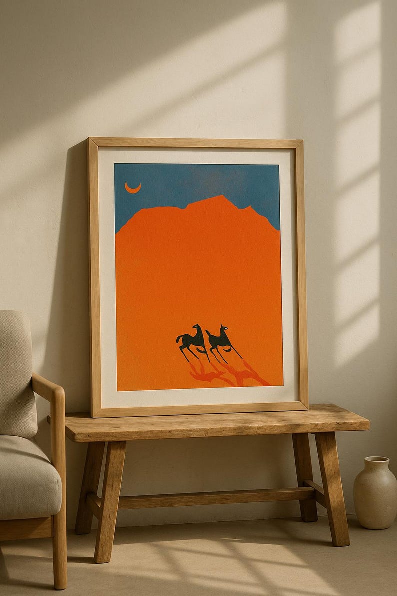 May include: Framed art print featuring a minimalist desert landscape. The artwork depicts two stylized black animals with long shadows against an orange background, with a blue sky and a crescent moon. The print is in a light wood frame.