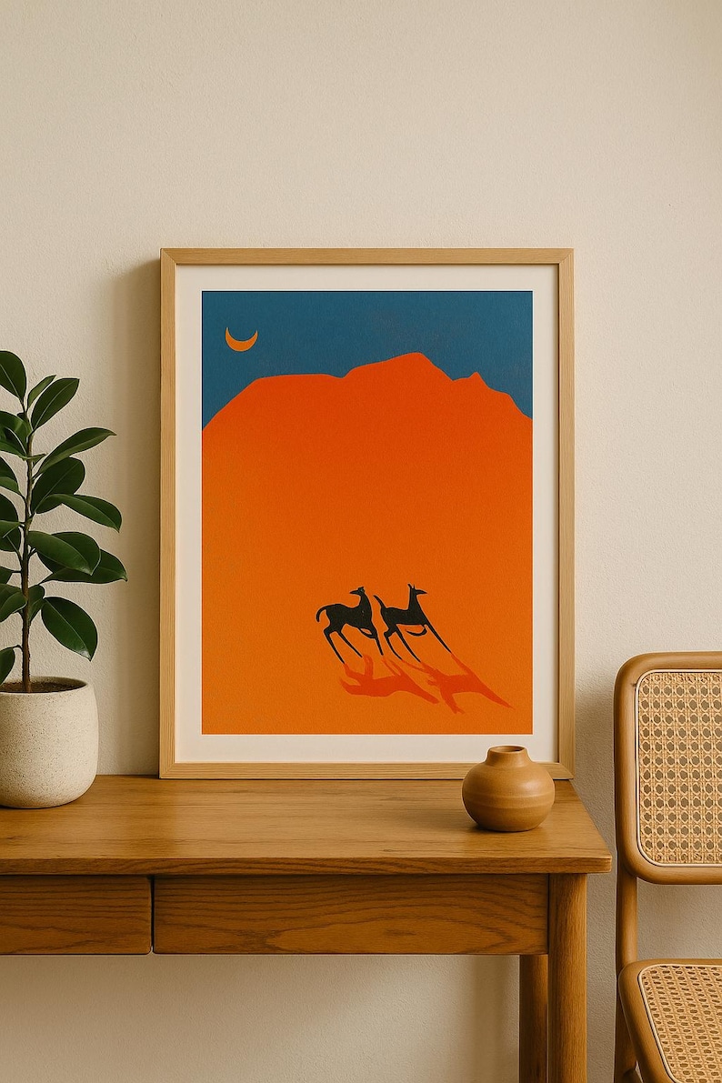 May include: Framed art print featuring a minimalist desert landscape. The artwork depicts two black animals running across an orange desert under a blue sky with a crescent moon. The print is in a wooden frame.