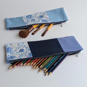 May include: Two fabric pencil cases with zippers. One is light blue with a floral pattern and holds makeup brushes. The other is navy blue with a floral pattern and holds coloured pencils. Both cases are on a white background.