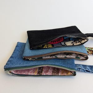 May include: Three denim pouches in varying shades of blue and black, each with a zipper closure. The interiors feature colorful patterned fabric. A black strap and a patterned strap are visible.