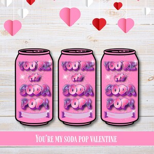 Op de afbeelding: Drie roze Valentijnsdagkaarten in de vorm van frisdrankblikjes met de tekst "YOU'RE MY SODA POP!" in paars en wit. De kaarten hebben onderaan een banner voor "From:". De zin "YOU'RE MY SODA POP VALENTINE" staat onderaan.