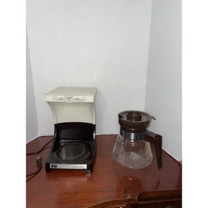 May include: A two-tone cream and dark brown electric coffee maker with a matching glass carafe. The machine features a cream-colored top lid with decorative scrollwork and a dark brown base. The glass carafe has a brown lid and handle.