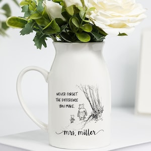 Puede incluir: Una jarra de cerámica blanca con asa, llena de rosas blancas y vegetación. La jarra tiene una ilustración en blanco y negro y el texto: "Never forget the difference you make. mrs. miller."