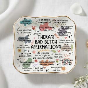 Könnte beinhalten: Eine weiße Keramik-Schmuckschale mit goldenem Rand, auf der "Thera's Bad Bitch Affirmations" und verschiedene ermächtigende Sätze stehen. Ein goldener Ring mit Diamant und weiße Blumen liegen daneben.