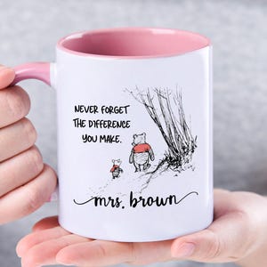 Könnte beinhalten: Weiße Keramik-Tasse mit rosa Innenraum und Henkel. Die Tasse zeigt eine Schwarz-Weiß-Illustration von Winnie Puuh und Ferkel, mit dem Text "NEVER FORGET THE DIFFERENCE YOU MAKE" und "mrs. brown" in schwarzer Schrift.