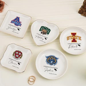 Puede incluir: Una colección de platos de cerámica blancos con bordes dorados, cada uno con un logotipo universitario y texto personalizado. Los platos tienen varias formas, incluyendo cuadrada, corazón y redonda, y están decorados con diseños temáticos de graduación.