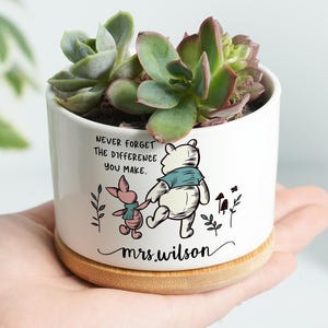 May include: White ceramic planter with a succulent plant, featuring Winnie the Pooh and Piglet illustrations. The planter has the text "Never forget the difference you make" and "mrs. wilson". The planter sits on a wooden base.