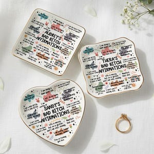May include: Three ceramic trinket dishes in square, rectangular, and heart shapes. Each dish has gold trim and affirmations in black, pink, and teal. The text on the dishes reads "Audrey's Bad Bitch Affirmations", "Thera's Bad Bitch Affirmations", and "Sandy's Bad Bitch Affirmations".