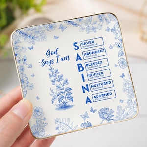Puede incluir: Plato cuadrado de cerámica con borde dorado, con un diseño floral azul y las palabras "Dios dice que soy." El plato incluye las palabras "Salvado, Abundante, Bendecido, Invitado, Nutrido, Adornado" con versículos bíblicos.