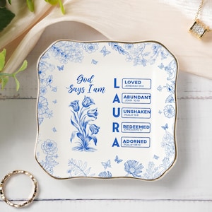 May include: A square, white ceramic trinket dish with a blue floral design and gold trim. The dish features the text "God Says I Am" and the name "LAURA" with associated affirmations. A gold ring and necklace are also visible.