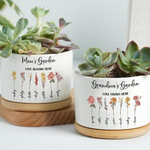 Op de afbeelding: Twee witte keramische plantenpotten met vetplanten, elk op een houten voet. Eén leest "Mom's Garden" en de andere "Grandma's Garden", beide met bloemenillustraties en de zin "Love Blooms Here".