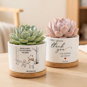 Pode incluir: Dois vasos cilíndricos de cerâmica branca com pratos de bambu, cada um contendo uma suculenta. Um apresenta uma ilustração do Ursinho Pooh com o texto "NEVER FORGET THE DIFFERENCE YOU MAKE" e "Mrs. Garcia". O outro diz "Mrs. Garcia thank you FOR HELPING ME GROW" com um coração vermelho e "Audrey".