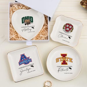 Puede incluir: Una colección de platos de cerámica personalizados de varias formas. Un plato en forma de corazón presenta el logotipo de la Universidad de Ohio, mientras que otros muestran emblemas universitarios y nombres. Cada plato tiene un borde dorado y se presenta en una caja de regalo blanca.
