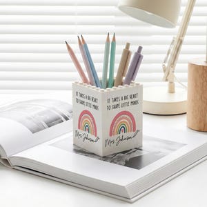 Op de afbeelding: Een witte vierkante pennenhouder gemaakt van in elkaar grijpende bouwstenen. De voorkant is voorzien van een kleurrijke regenboogafbeelding, een klein hartje en de gedrukte tekst: It takes a big heart to shape little minds, gevolgd door Mrs. Johnson.