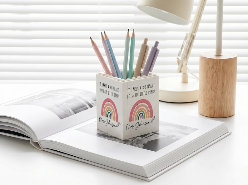 Op de afbeelding: Een witte vierkante pennenhouder gemaakt van in elkaar grijpende bouwstenen. De voorkant is voorzien van een kleurrijke regenboogafbeelding, een klein hartje en de gedrukte tekst: It takes a big heart to shape little minds, gevolgd door Mrs. Johnson.