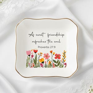 Puede incluir: Plato de cerámica blanco con borde dorado y diseño floral. El plato presenta el texto "A sweet friendship refreshes the soul" y Proverbios 27:9. El diseño floral incluye flores rosas, rojas, amarillas y naranjas.