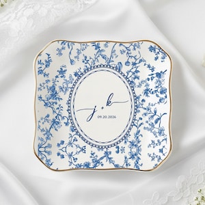 Pode incluir: Prato quadrado de cerâmica com um padrão floral azul e borda dourada. O centro apresenta um oval com as iniciais "j + k" e a data "09.20.2026" em escrita azul marinho. O prato está sobre uma superfície acetinada branca.