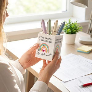 Op de afbeelding: Een witte vierkante pennenhouder gemaakt van bouwstenen, voorzien van een kleurrijke regenboogafbeelding, een klein rood hartje en de gedrukte tekst "It takes a big heart to shape little minds" samen met de naam "Mrs. Anderson" in zwart schrift.