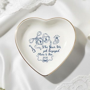 May include: A heart-shaped ceramic ring dish with a gold rim. The white dish has a blue illustration of two lockets, a ring, and a champagne bottle. Text reads "The Year We got Engaged Olivia & Ben 2025".