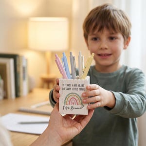 Op de afbeelding: Een witte vierkante pennenhouder met een kleurrijke regenboogafbeelding, de tekst "It takes a big heart to shape little minds" en de naam "Mrs. Brown" op de voorzijde, gevuld met vijf pastelkleurige pennen in roze, blauw, grijs, wit en geel.