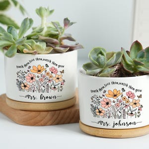 Pode incluir: Dois vasos de cerâmica brancos com suculentas, cada um com um design floral e o texto "teach them, love them, watch them grow". Um vaso é rotulado "Mrs. Brown", o outro "Mrs. Johnson". Ambos estão em pires de madeira.