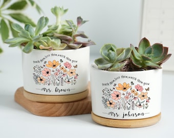 May include: Two white ceramic pots with succulents, each featuring a floral design and the text "teach them, love them, watch them grow." One pot is labeled "Mrs. Brown," the other "Mrs. Johnson." Both sit on wooden saucers.