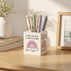 Op de afbeelding: Een witte vierkante pennenhouder gemaakt van bouwstenen, voorzien van de tekst 'It takes a big heart to shape little minds', een kleurrijke regenboogafbeelding, de naam 'Mrs. Taylor' met een potloodicoon, gevuld met vier pastelkleurige pennen.