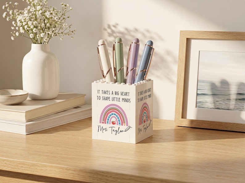 Op de afbeelding: Een witte vierkante pennenhouder gemaakt van bouwstenen, voorzien van de tekst 'It takes a big heart to shape little minds', een kleurrijke regenboogafbeelding, de naam 'Mrs. Taylor' met een potloodicoon, gevuld met vier pastelkleurige pennen.