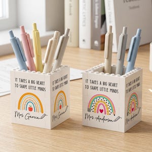 Op de afbeelding: Twee vierkante pennenbakjes gemaakt van in elkaar grijpende bouwstenen. Elke houder bevat de tekst "It takes a big heart to shape little minds" boven een kleurrijke regenboogafbeelding en een gepersonaliseerde naam, Mrs. Garcia of Mrs. Anderson.