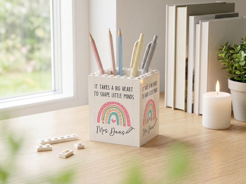 Op de afbeelding: Een witte vierkante pennenhouder gemaakt van in elkaar grijpende bouwstenen. De voorkant is voorzien van een kleurrijke regenboogafbeelding, de tekst "IT TAKES A BIG HEART TO SHAPE LITTLE MINDS" en de naam "Mrs. Davis" in zwart schrift.