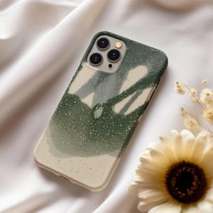 May include: A green and cream-colored phone case with a speckled, abstract design. The case has a matte finish and a raised edge around the camera lenses. The design features a blend of green and cream tones, creating a unique aesthetic.