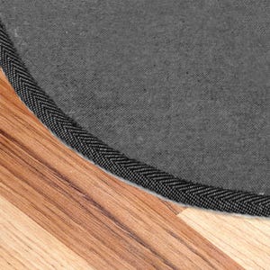 May include: Close-up of the edge of a round gray rug with a black stitched border. The rug is on a wood floor.