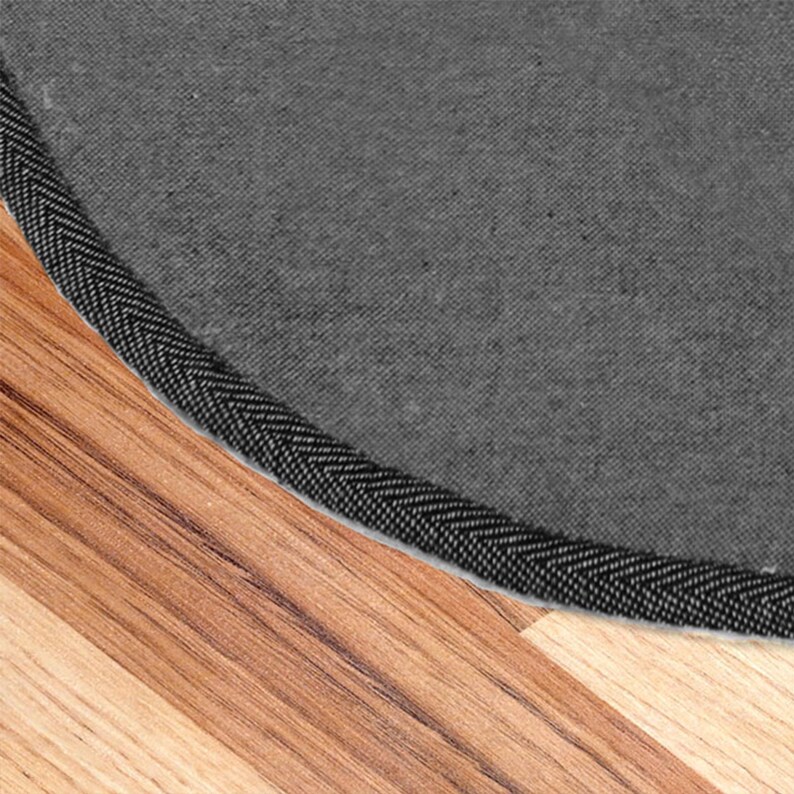 May include: Close-up of the edge of a round gray rug with a black stitched border. The rug is on a wood floor.