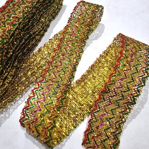 May include: Gold metallic ribbon with a zigzag pattern, featuring red, green, and purple accents. The ribbon is displayed in a roll and in sections, showcasing its texture and design. Ideal for crafting and decorative projects.
