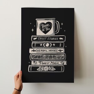 May include: Black and white art print showing a stack of books with titles such as "Ghost Stories" and "Forest Magic." A mug with a heart and the words "Books & Tea" sits on top. The print is held by a person.
