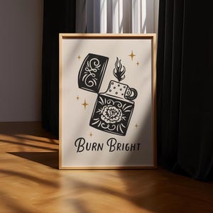 以下が含まれることがあります： フレーム入りアートプリント。黒と白のイラストで、花柄のデザインと「Burn Bright」の文字が入った開いたライターが描かれています。金色の星のアクセントがデザインを飾っています。