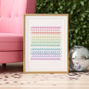 Puede incluir: Una impresión artística enmarcada con líneas coloridas y estampadas en tonos arcoíris. La obra de arte se exhibe sobre un suelo de madera, con una silla rosa y una bola de discoteca en el fondo.