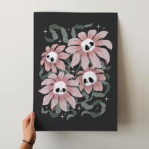 May include: A dark gray art print featuring a floral design with a twist. The flowers are pink with white skull centers, surrounded by green leaves and sparkling stars. The artwork is held by a person.