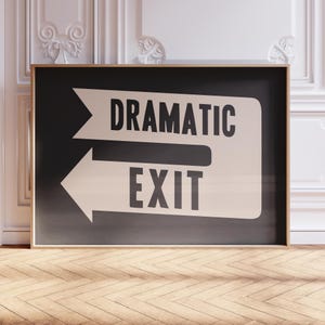 Puede incluir: Un cuadro enmarcado con las palabras "DRAMATIC EXIT" en negrita y mayúsculas negras. Las palabras están sobre un fondo negro con una flecha blanca que apunta hacia la izquierda. El marco es de color madera clara.