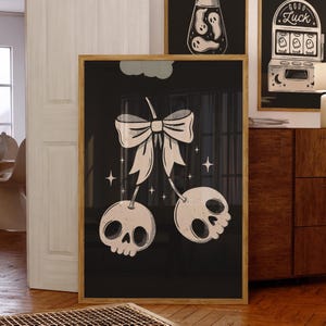 May include: A framed art print with a black background, depicting a white bow and two skull-shaped cherries. The skulls have eye sockets and a mouth, with star accents. The artwork is in a wooden frame.