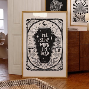 May include: Black and white illustration of a coffin with the text "I'll sleep when I'm dead" inside. The coffin is surrounded by a decorative frame with stars, spiderwebs, and other gothic elements.