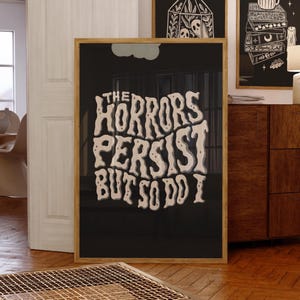 以下が含まれることがあります： 「THE HORRORS PERSIST BUT SO DO I」のテキストが、黒い背景にヴィンテージ風のオフホワイトのフォントで描かれた額入りのアートプリント。フレームは明るい木の色です。他の額入りのアート作品と一緒に部屋に飾られています。