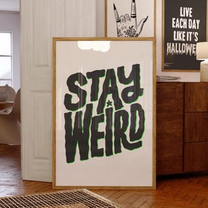 Peut inclure: Affiche encadrée avec les mots "STAY WEIRD" en grandes lettres noires avec un contour vert. L'affiche est affichée sur un fond blanc et possède un cadre en bois. D'autres œuvres encadrées sont visibles en arrière-plan.
