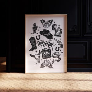 May include: Framed black and white print with Western-themed illustrations. The design includes a cowboy hat, boots, a revolver, a snake, a skull, a cactus, and the word "HOWDY" on a banner. The print is in a wooden frame.
