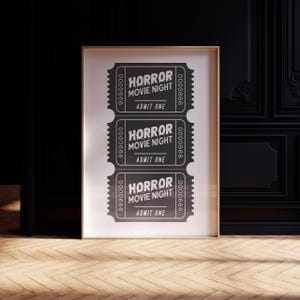 May include: A framed print displaying three black and white cinema tickets. Each ticket is printed with "HORROR MOVIE NIGHT" and "ADMIT ONE", along with a serial number. The print is in a light wood frame, set against a dark backdrop.